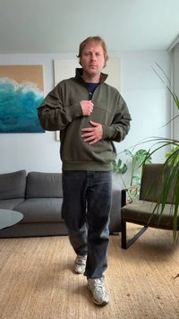 Olivovo zelený pulóver s polovičným zipsom, rebrovaným golierom a manžetami; nosený cez tmavú košeľu a kombinovaný s tmavými džínsami a teniskami.