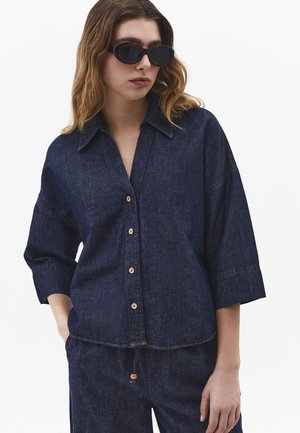 Kvinde iført mørk denim skjorte med knapper og trekvartlange ærmer samt matchende bukser, accessoriseret med sorte ovale solbriller.