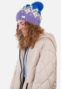 Purple knitted beanie with a blue pom-pom, featuring a multicolored pattern of green and blue speckles. Tag visible. Worn with a light jacket.