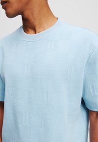 Lyseblå strikket sweater med et tekstureret ternet mønster og diskret logo-detaillering, med rund halsudskæring og korte ærmer.