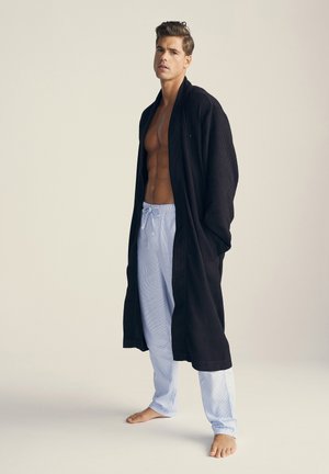 Sort morgenkåbe med vaffelmønster og sjalskrave, båret over lyseblå stribede pyjamasbukser. Modellen står barfodet og viser outfittet.