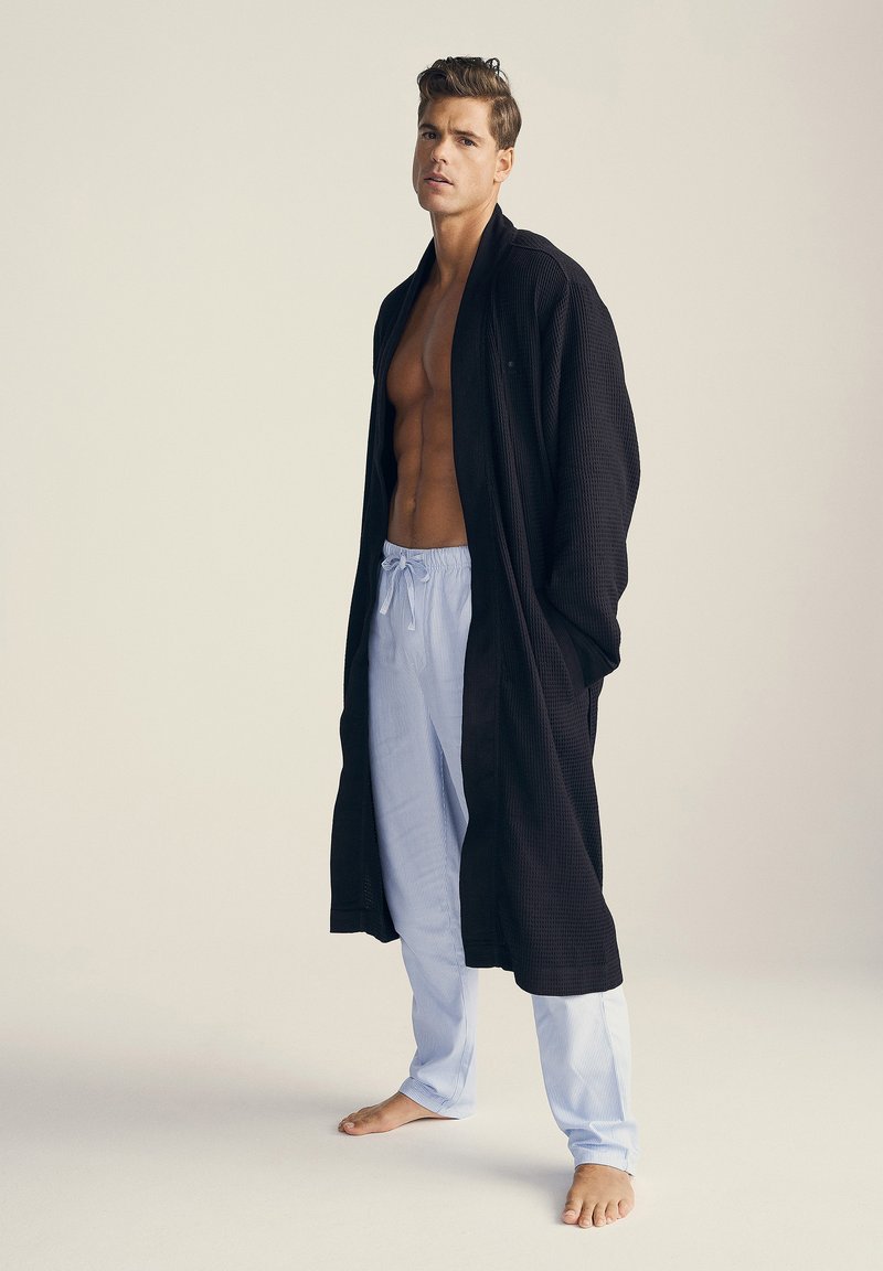 Black waffle-pattern robe with a shawl collar, worn over light blue striped pajama pants. Model stands barefoot, showcasing the outfit.