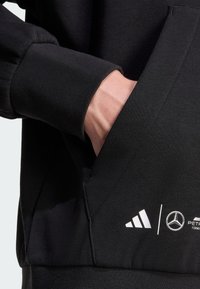 Sort sweatshirt med struktureret stof, ribstrikkede manchetter og en frontlomme med hvide Adidas- og Mercedes-logoer.