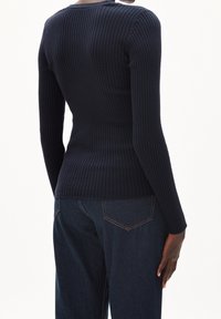 Tmavě modrý žebrovaný top s dlouhými rukávy a kulatým výstřihem, přiléhavý střih a texturovaná látka; v kombinaci s tmavými džínovými kalhotami s kapsami na zadní straně.