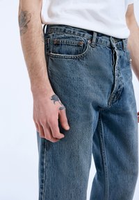 Person iført blå denimjeans og en hvid skjorte, med tatoveringer af en rose på underarmen og hånden.