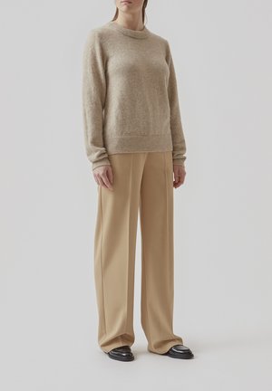 Beige uldtrøje med rund halsudskæring og ribbede manchetter, parret med lys beige bukser med vidde og sorte sko.