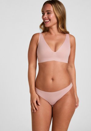Femme souriante portant un soutien-gorge sans coutures rose clair et un slip taille basse assorti, debout devant un fond gris uni, regardant vers la droite.