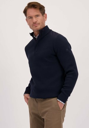 Man met lichtbruin haar, gekleed in een marineblauwe quarter-zip trui over een gemusterd shirt en kaki broek, staand met zijn handen in zijn zakken tegen een effen achtergrond.