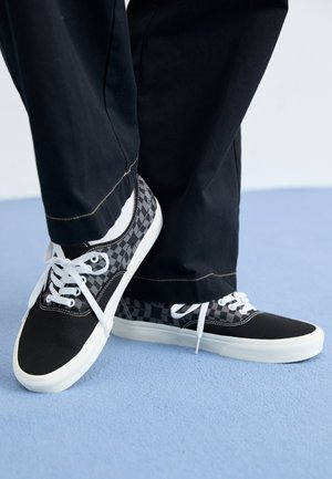 Čierne plátkové tenisky s šedým kockovaným vzorom, bielymi šnúrkami a gumovými podrážkami, skombinované s čiernymi nohavicami na modrom povrchu.