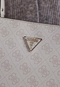 Beige handväska med ett texturerat mönster, med en framträdande guldig triangel-logotyp på framsidan och ett robust vitt handtag.