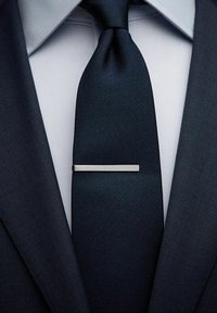 Marinblå slips med texturerat mönster, fäst med en stilren silverfärgad slipsklämma. Bakgrunden består av en krispigt vit skjorta och en mörk kostymjacka.
