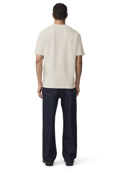 Mężczyzna stojący tyłem, ubrany w prosty beżowy t-shirt z krótkim rękawem oraz ciemnoniebieskie, proste dżinsy i czarne buty.