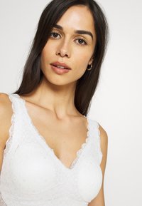 Bijeli čipkani bralette s V-izrezom, valovitim rubovima i teksturiranim kružnim uzorcima. Meki materijal s podesivim trakama.