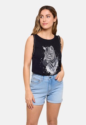 Mujer con una camiseta negra sin mangas con gráfico de cebra y pantalones cortos de mezclilla azul claro remangados, posando con una mano en el bolsillo.