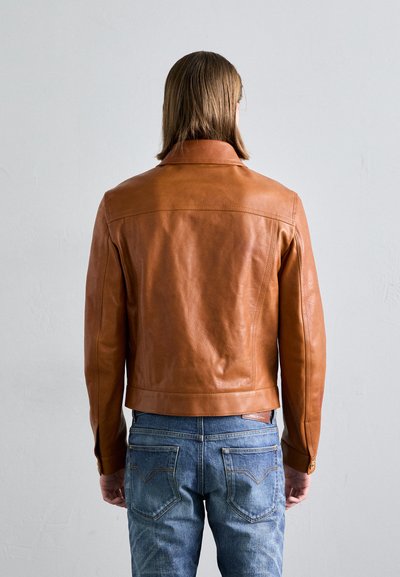 Braune Lederjacke mit Kragen, taillierte Form und glatter Textur. Kombiniert mit blauen Jeans mit gestickten Gesäßtaschen.