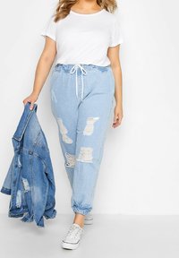 Joggers de mezclilla azul claro desgastados con puños elásticos y cintura con cordón, combinados con una camiseta blanca y una chaqueta de mezclilla. Zapatillas blancas.