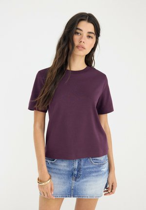 Jeune femme aux longs cheveux foncés portant un t-shirt violet à manches courtes et une jupe en jean bleue sur un fond clair uni.