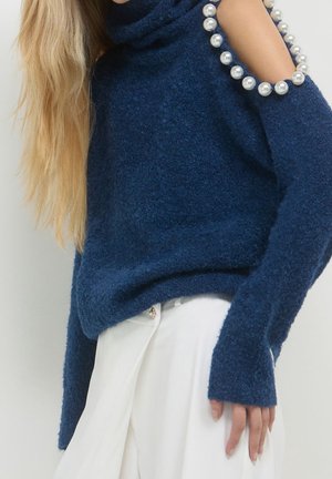 Pull bleu duveteux avec détail d'épaule dénudée orné de perles, porté avec un pantalon blanc taille haute.