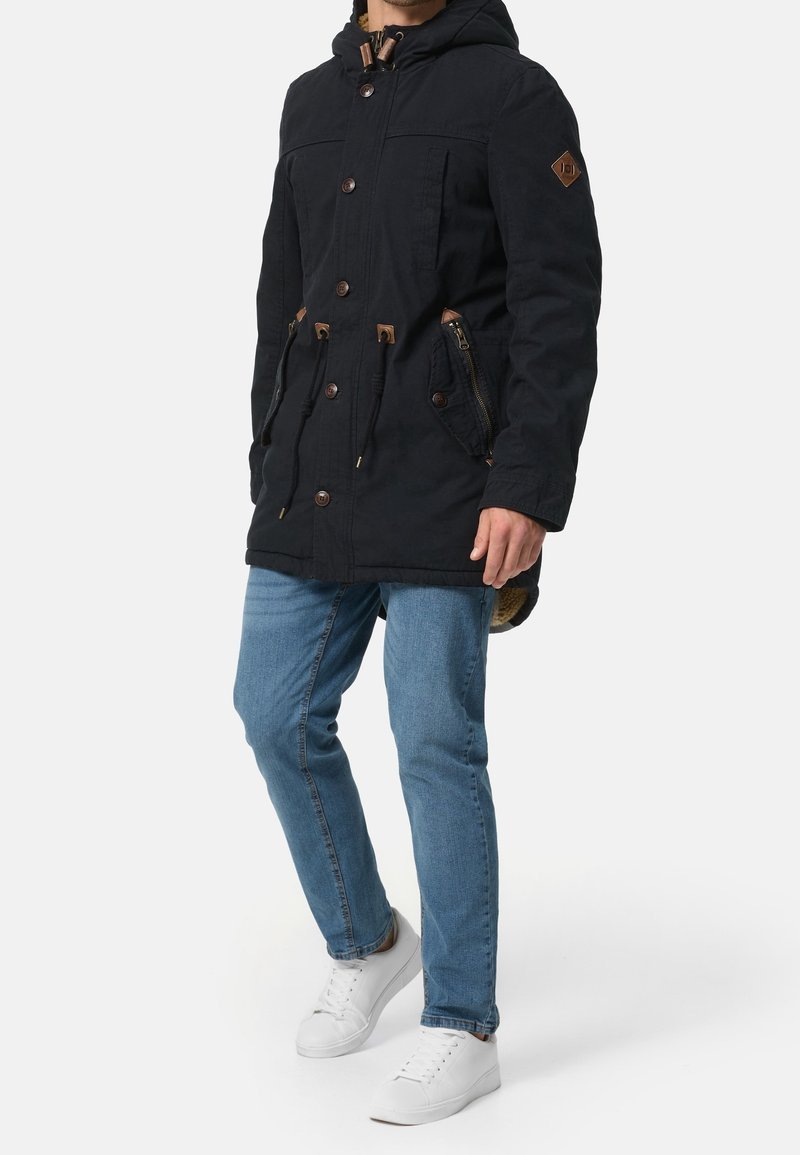 Parka bleu marine avec capuche, cordons de serrage et plusieurs poches. Comprend des accents de quincaillerie marron et une doublure intérieure matelassée.