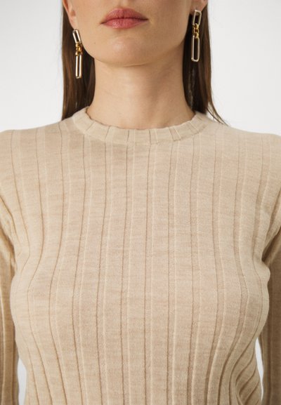 Sweater en tricot côtelé beige avec un col rond, présentant des lignes verticales et une texture douce. Des boucles d'oreilles en or ajoutent une touche métallique.