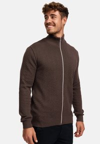 Brauner Reißverschluss-Pullover mit strukturiertem Fischgrätmuster, geripptem Kragen und Bündchen sowie einem glatten Reißverschlussdetail vorne.