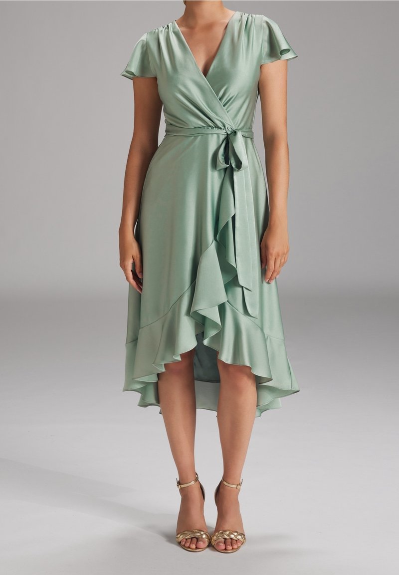 Femme portant une robe portefeuille en satin vert clair avec de courtes manches volantées et un ourlet haut-bas à volants, associée à des talons à lanières dorés.