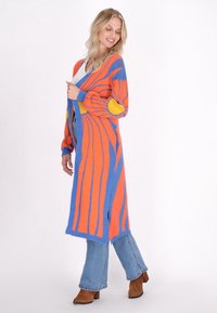 Lungo cardigan lavorato a maglia in arancione, blu e giallo. Presenta un motivo audace, polsini a costine e fessure laterali per facilitare il movimento.