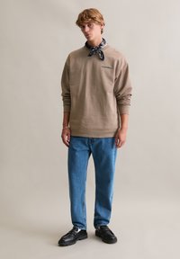 Beżowa bluza z długim rękawem i okrągłym dekoltem z haftowanym detalem, zestawiona z jasno-niebieskimi dżinsami i czarnymi butami wsuwanymi. Akcent bandany.