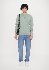 Felpa verde a maniche lunghe con logo EA7 sul petto, abbinata a jeans azzurri e sneaker bianche. Borsa a spalla nera con hardware argentato.