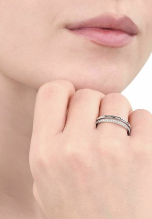 Nahaufnahme einer Hand mit einem silbernen Ring mit kleinen Diamanten am Ringfinger, die sich in der Nähe eines neutral gefärbten Gesichts und der Lippen befindet.