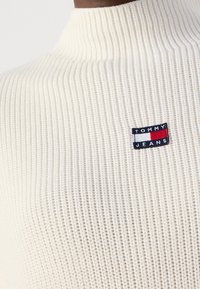 Pull à col roulé en maille côtelée crème avec petit logo rectangulaire Tommy Jeans en rouge, blanc et marine sur la poitrine.