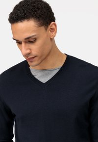 Marineblauwe V-hals gebreide trui met een gestructureerde afwerking, gedragen over een grijze ronde hals t-shirt. Toont subtiele stikdetails. Binnenste kraag zichtbaar.