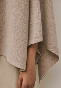 Poncho de lana beige, textura suave, caída amplia, con dobladillo acanalado y diseño sin costuras. Mano parcialmente cubriendo el lado inferior.