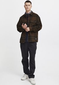 Chemise à carreaux marron avec une poche poitrine, portée sur une chemise sombre et associée à un pantalon sombre et des baskets blanches.