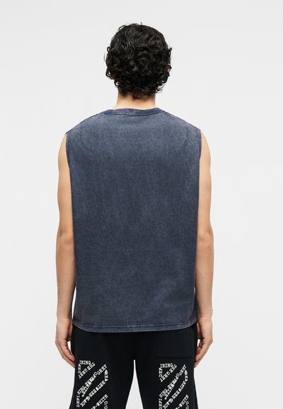 Άνδρας με σγουρά σκούρα μαλλιά, φορώντας χωρίς μανίκια ξεθωριασμένο σκούρο γκρι πουκάμισο και μαύρο παντελόνι με λευκό κείμενο, στέκεται πλάτη σε λευκό φόντο.