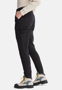 Personne portant un pantalon cargo noir avec plusieurs poches et des chaussures de randonnée beiges avec des accents bleus et des lacets jaunes sur un fond blanc.