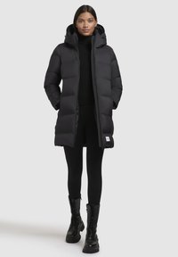 Zwarte pufferjas met capuchon, met een doorgestikt ontwerp, rits aan de voorkant en zijzakken. Draagt over een zwarte top en legging, gecombineerd met zwarte laarzen.