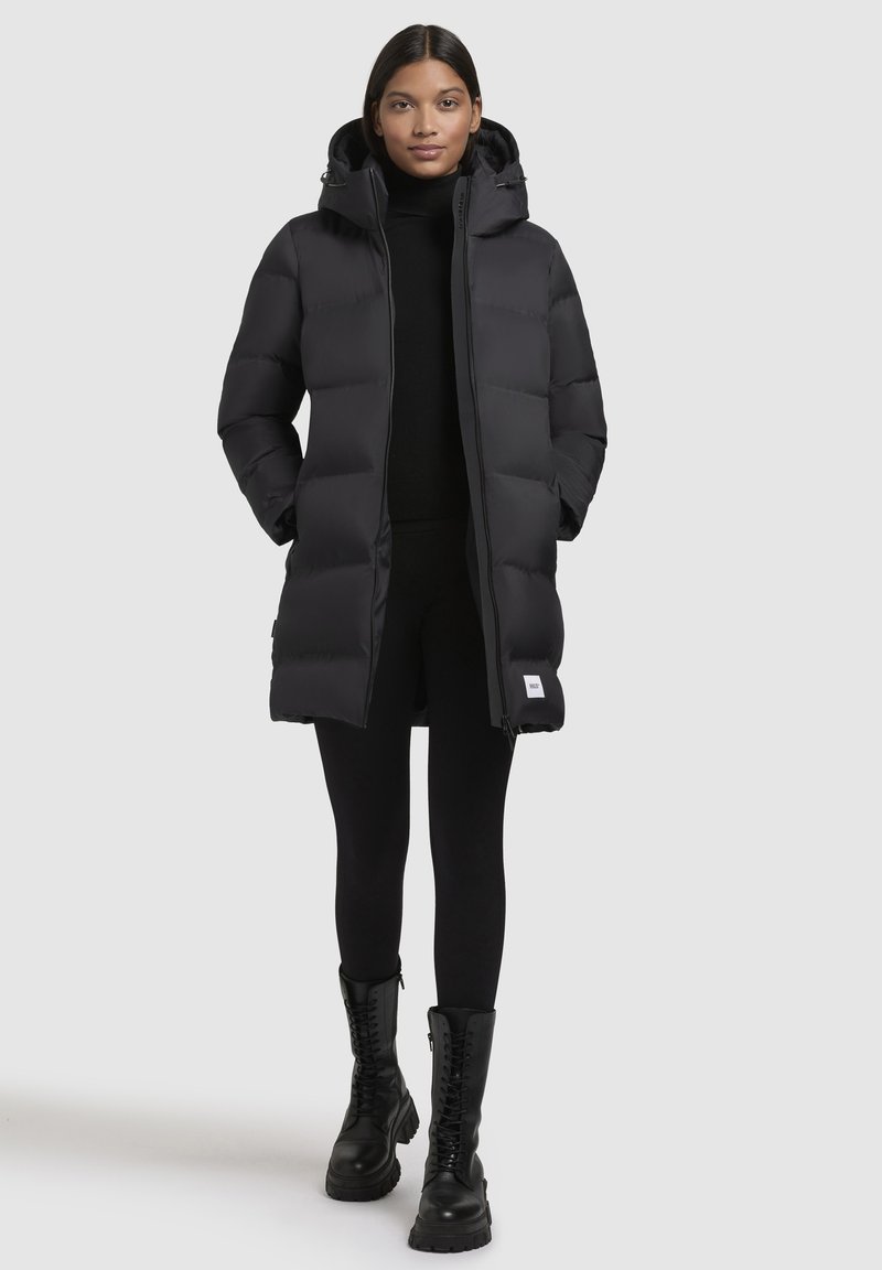 Zwarte pufferjas met capuchon, met een doorgestikt ontwerp, rits aan de voorkant en zijzakken. Draagt over een zwarte top en legging, gecombineerd met zwarte laarzen.