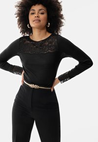 Zwarte longsleeve top met kantdetails op de schouders en mouwen, gecombineerd met op maat gemaakte zwarte broek en een riem met gouden accenten.
