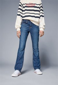 Person med vita sneakers, blå jeans och en vit och svart randig Tommy Hilfiger sweatshirt mot en enkel grå bakgrund.