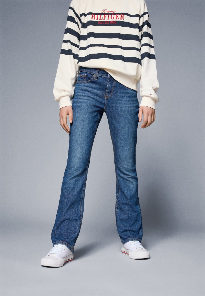 Person med vita sneakers, blå jeans och en vit och svart randig Tommy Hilfiger sweatshirt mot en enkel grå bakgrund.