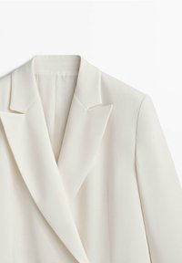 Veste blanche sur mesure avec larges revers et épaules structurées, présentée sur un fond blanc.