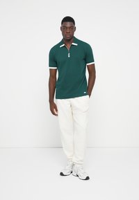 Grøn polo-shirt med lynlåskrave, hvide striber på ærmerne, båret med hvide sweatpants og grå træningssko. Enkelts design og teksturer.