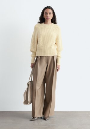 Femme portant un pull côtelé couleur crème, un pantalon large beige, des chaussures pointues grises, portant un sac cabas beige, debout contre un mur blanc.