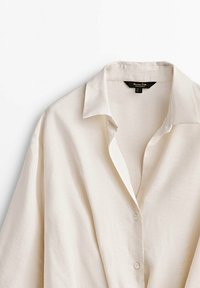 Licht beige knoopshirt met een kraag, zachte textuur en een relaxed pasvorm. Bevat een label dat aan de binnenkant van de kraag is genaaid.