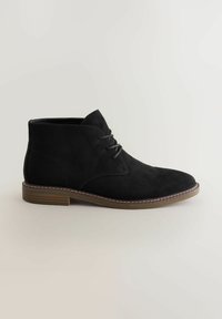 Sorte suede ankelstøvler med rund tå og snørebånd, med en brun sål og syninger, sat op mod en lys baggrund.