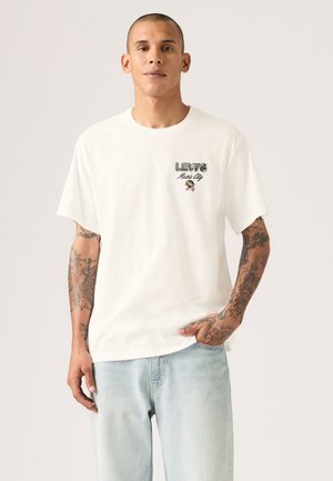 Hombre con tatuajes en ambos brazos lleva una camiseta blanca de Levi's con el texto "Music City" y vaqueros azul claro sobre un fondo liso.