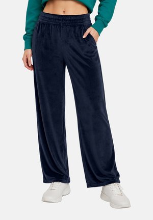 Femme portant un pantalon large en velours bleu marine avec taille élastique et baskets blanches, main dans la poche, top court vert visible.