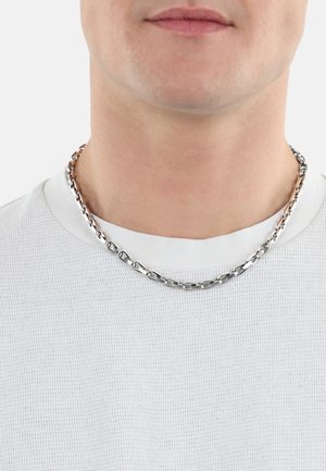 Collana a catena in tono argento con anelli rettangolari intrecciati, indossata sopra una camicia bianca a trama, vicino al collo.