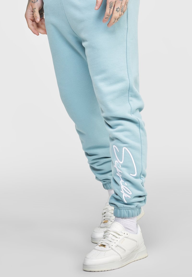 Světle modré teplákové kalhoty s tapered nohavicí, se bílým písmem na boku a elastickými manžetami, doplněné bílými teniskami.